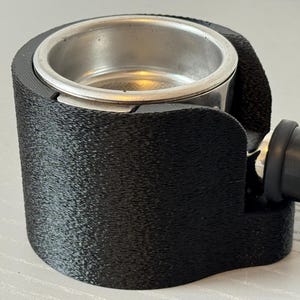 Könnte beinhalten: Ein schwarzer Siebträgerhalter für eine Espressomaschine mit einem Edelstahl-Filterkorb. Der Halter besteht aus einem strukturierten, mattschwarzen Material. Der Griff ist schwarz mit einem Gummiüberzug.