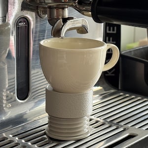 Peut inclure: Une tasse à expresso en céramique couleur crème est posée sur un support cylindrique strié gris clair. La tasse est placée directement sous le porte-filtre métallique d'une machine à expresso en acier inoxydable avec une grille d'égouttage.