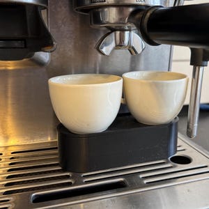 Könnte beinhalten: Zwei cremefarbene Espressotassen aus Keramik stehen auf einer schwarzen Plattform unter dem Auslauf einer Espressomaschine aus Edelstahl. Die Tassen sind klein, haben eine runde Form und einen Henkel. Die Maschine hat ein silbernes Finish.