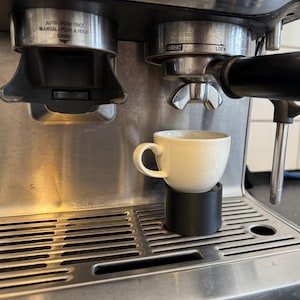 Op de afbeelding: Een roestvrijstalen espressomachine met een witte keramische beker op een zwarte basis. De machine heeft een portafilter en een stoompijp. De beker staat onder de portafilter.