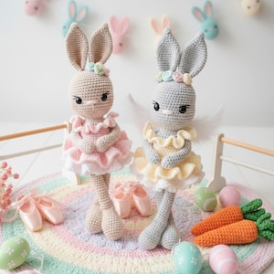 Może przedstawiać: Dwie szydełkowe lalki zajączki ubrane jako baletnice, jedna w różu, druga w żółci, stoją na kolorowym dywanie. Mają kwiatowe korony i skrzydła baletowe. Baletki, marchewki i pisanki je otaczają, z głowami królików na ścianie.