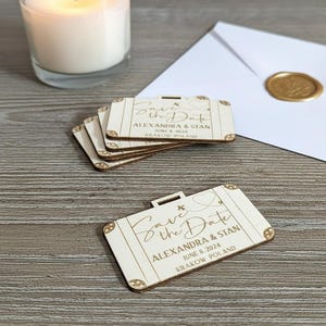 Puede incluir: Recuerdos de boda de madera con forma de maleta con la inscripci&oacute;n "Save the Date", nombres, fecha y lugar. Una vela encendida y un sobre sellado est&aacute;n en el fondo. Los recuerdos est&aacute;n hechos de madera clara.