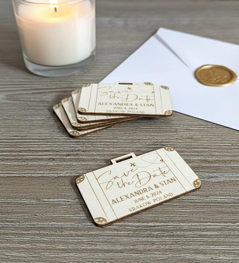 Puede incluir: Recuerdos de boda de madera con forma de maleta con la inscripci&oacute;n "Save the Date", nombres, fecha y lugar. Una vela encendida y un sobre sellado est&aacute;n en el fondo. Los recuerdos est&aacute;n hechos de madera clara.