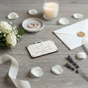 Puede incluir: Una composici&oacute;n de boda con una tarjeta de invitaci&oacute;n en forma de maleta de madera con el texto "Save the Date". Incluye un sobre blanco con un sello dorado, anillos de boda, una vela encendida, un ramo de rosas blancas y p&eacute;talos esparcidos.