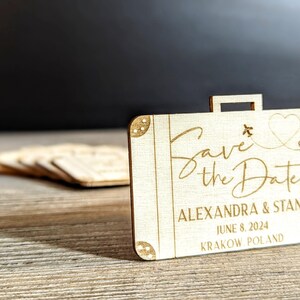 Puede incluir: Recuerdo de boda de madera en forma de maleta con la frase "Save the Date" en escritura cursiva. Incluye un coraz&oacute;n y un avi&oacute;n. Los nombres Alexandra & Stan, la fecha y el lugar est&aacute;n grabados.