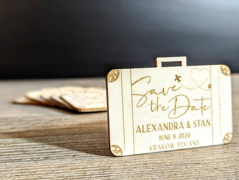 Puede incluir: Recuerdo de boda de madera en forma de maleta con la frase "Save the Date" en escritura cursiva. Incluye un coraz&oacute;n y un avi&oacute;n. Los nombres Alexandra & Stan, la fecha y el lugar est&aacute;n grabados.