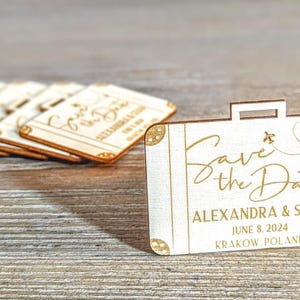 Puede incluir: Recuerdos de boda de madera en forma de maleta con el texto "Save the Date" y los nombres Alexandra & Stan, 8 de junio de 2024, Cracovia, Polonia. Los recuerdos son de madera clara con detalles dorados.