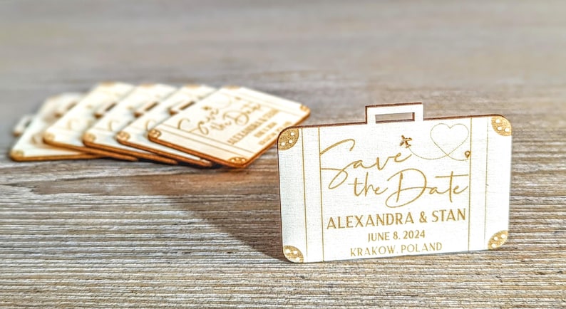 Puede incluir: Recuerdos de boda de madera en forma de maleta con el texto "Save the Date" y los nombres Alexandra & Stan, 8 de junio de 2024, Cracovia, Polonia. Los recuerdos son de madera clara con detalles dorados.