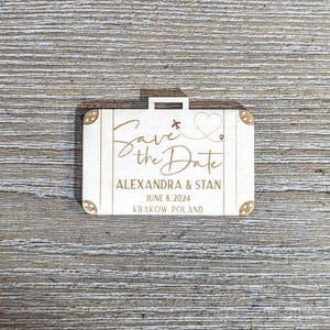 Puede incluir: Un anuncio de boda de madera con forma de maleta con las palabras "Save the Date" en escritura elegante. Tambi&eacute;n se muestran los nombres "Alexandra & Stan", la fecha "8 de junio de 2024" y la ubicaci&oacute;n "Krakow, Polonia".