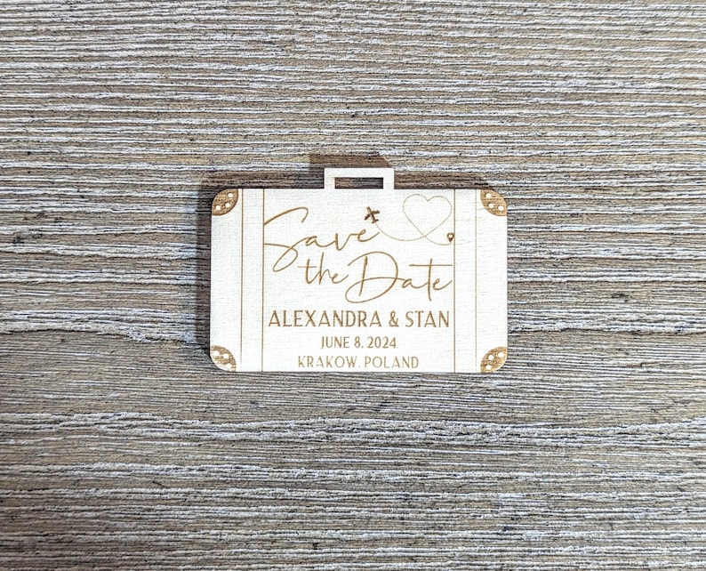 Puede incluir: Un anuncio de boda de madera con forma de maleta con las palabras "Save the Date" en escritura elegante. Tambi&eacute;n se muestran los nombres "Alexandra & Stan", la fecha "8 de junio de 2024" y la ubicaci&oacute;n "Krakow, Polonia".