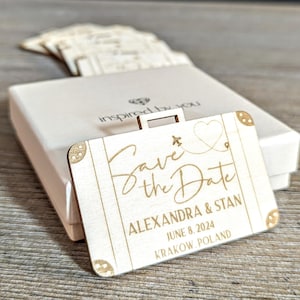Puede incluir: Un recuerdo de boda en forma de maleta de madera clara con la inscripci&oacute;n "Save the Date", los nombres ALEXANDRA & STAN, la fecha 8 de junio de 2024 y la ubicaci&oacute;n Cracovia, Polonia. Se encuentra frente a una caja blanca.