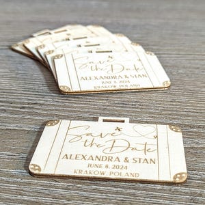 Puede incluir: Peque&ntilde;os recuerdos de boda de madera en forma de maleta, grabados con "Save the Date", nombres, fecha y lugar. La fecha es el 8 de junio de 2024. Se ven detalles decorativos en las esquinas.