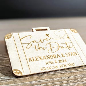 Puede incluir: Un anuncio de boda de madera con forma de maleta con las palabras "Save the Date" en escritura elegante. El anuncio incluye los nombres Alexandra & Stan, la fecha 8 de junio de 2024 y la ubicaci&oacute;n Cracovia, Polonia.
