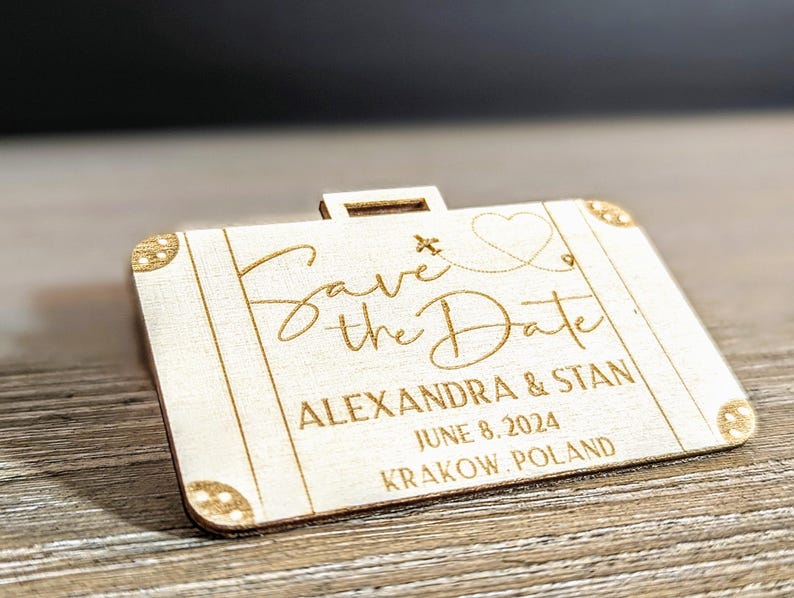 Puede incluir: Un anuncio de boda de madera con forma de maleta con las palabras "Save the Date" en escritura elegante. El anuncio incluye los nombres Alexandra & Stan, la fecha 8 de junio de 2024 y la ubicaci&oacute;n Cracovia, Polonia.