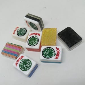 Może przedstawiać: Zbiór kafelków do gry w Mahjong, w tym kilka oznaczonych napisem "JOKER". Kafelki mają różne kolory i wzory, w tym zielone grafiki kołowe, złoty brokat i wzory w paski. Kafelki są prostokątne.