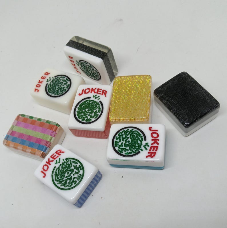 May include: A collection of Mahjong tiles, including several marked "JOKER". The tiles feature a variety of colors and designs, including green circular graphics, gold glitter, and striped patterns. The tiles are rectangular and have rounded edges.