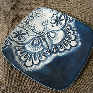 Puede incluir: Un plato de cerámica azul con un diseño de encaje blanco. El diseño de encaje tiene la forma de un búho.