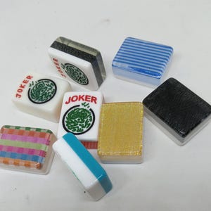 May include: A collection of colorful Mahjong tiles. The tiles feature various designs, including stripes, a gold glitter texture, and the word "JOKER" with a green dragon symbol. The tiles are square and have a glossy finish.