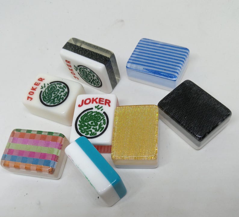 May include: A collection of colorful Mahjong tiles. The tiles feature various designs, including stripes, a gold glitter texture, and the word "JOKER" with a green dragon symbol. The tiles are square and have a glossy finish.