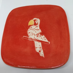 May include: A red ceramic square plate featuring a white parrot design. The parrot has a yellow beak and is perched on a branch.