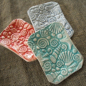May include: Three ceramic dish soap holders with a seashell and swirl design. The dishes are in shades of blue, teal, and coral.