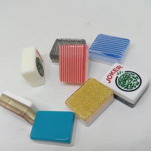 May include: A collection of colorful Mahjong tiles. The tiles feature various designs, including stripes, a green circular pattern, and the word "JOKER". Colors include red, blue, gold, and white. The tiles are arranged in a scattered pattern.