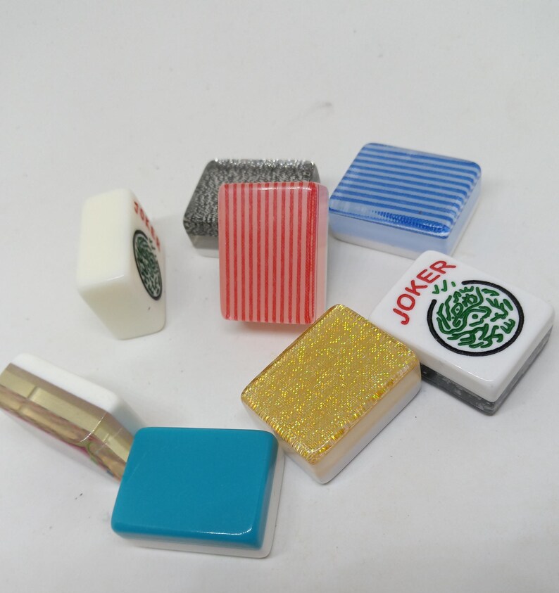 May include: A collection of colorful Mahjong tiles. The tiles feature various designs, including stripes, a green circular pattern, and the word "JOKER". Colors include red, blue, gold, and white. The tiles are arranged in a scattered pattern.