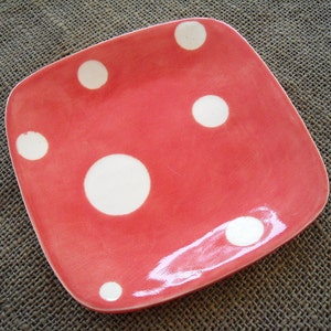 May include: A square, red ceramic plate with rounded corners and large, white polka dots. The plate has a glossy finish and is likely handmade. This decorative plate is suitable for serving food.