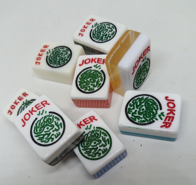 May include: A collection of white Mahjong tiles with green and black circular designs. Each tile has the word "JOKER" in red. Some tiles have colored edges, including pink, blue, and orange.