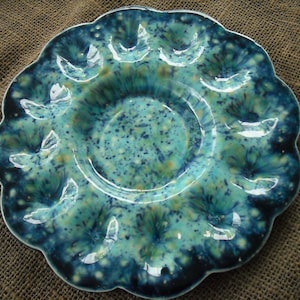 May include: A blue and green ceramic egg plate with a scalloped edge. The plate has a speckled glaze and is designed to hold six eggs.