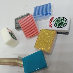 May include: A collection of colorful Mahjong tiles. The tiles feature various designs, including stripes, glitter, and a green circular pattern. One tile has the word "JOKER" printed on it. The tiles are in shades of blue, red, gold, and white.