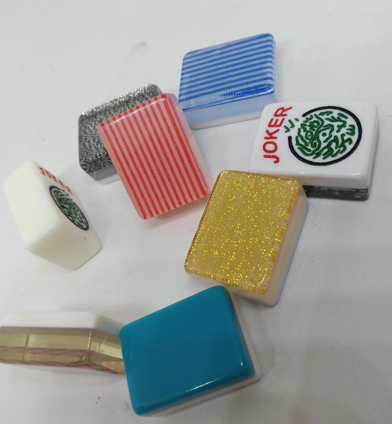 May include: A collection of colorful Mahjong tiles. The tiles feature various designs, including stripes, glitter, and a green circular pattern. One tile has the word "JOKER" printed on it. The tiles are in shades of blue, red, gold, and white.