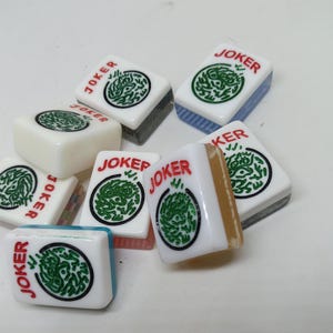 May include: A collection of Mahjong tiles, each displaying the word "JOKER" in red. The tiles are white with a green circular design. Some tiles have blue, gray, or tan edges. The tiles are scattered on a white surface.