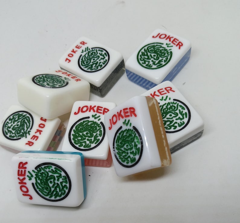 May include: A collection of Mahjong tiles, each displaying the word "JOKER" in red. The tiles are white with a green circular design. Some tiles have blue, gray, or tan edges. The tiles are scattered on a white surface.