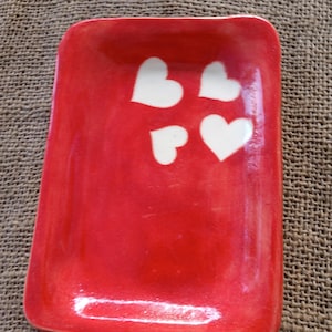 Puede incluir: Un plato rectangular de cerámica roja con cuatro diseños de corazones blancos. El plato tiene esquinas redondeadas y un acabado brillante. El fondo es una tela marrón texturizada.