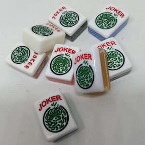 May include: A collection of white Mahjong tiles with the word "JOKER" in red. Each tile features a green circular design. The tiles have various colored edges, including blue, gray, and pink. The tiles are scattered on a white surface.