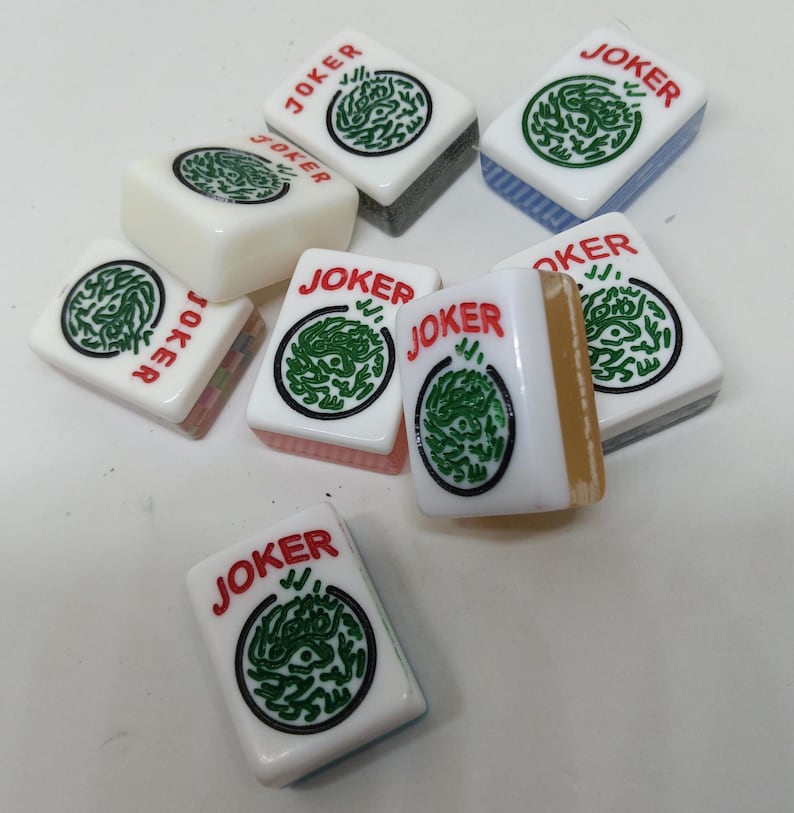 May include: A collection of white Mahjong tiles with the word "JOKER" in red. Each tile features a green circular design. The tiles have various colored edges, including blue, gray, and pink. The tiles are scattered on a white surface.