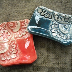 May include: Two ceramic soap dishes with a white floral design. One dish is red and the other is blue.