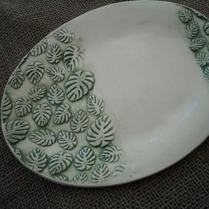 May include: Oval, off-white ceramic serving platter with a raised, green leaf design. The leaves are arranged around the edge of the platter. The platter is approximately 12 inches long and 9 inches wide.