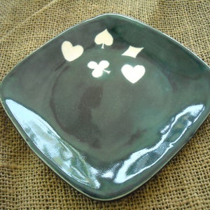 May include: A square, dark teal ceramic plate with rounded corners. The plate features white playing card suit symbols: hearts, spades, clubs, and diamonds. The plate has a glossy finish and is set against a textured background.