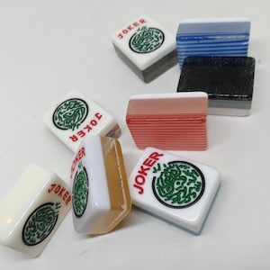 May include: A collection of colorful Mahjong tiles. Several tiles feature the word "JOKER" in red, along with a green circular design. Other tiles are solid colors, including blue, black, and red. The tiles are arranged on a white surface.