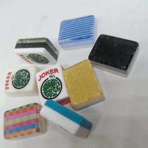 May include: A collection of colorful Mahjong tiles. The tiles feature various designs, including stripes, solid colors, and the word "JOKER" with a green dragon symbol. The tiles are in shades of blue, gold, black, and white.