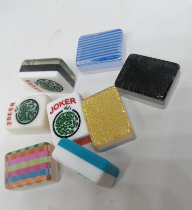 May include: A collection of colorful Mahjong tiles. The tiles feature various designs, including stripes, solid colors, and the word "JOKER" with a green dragon symbol. The tiles are in shades of blue, gold, black, and white.