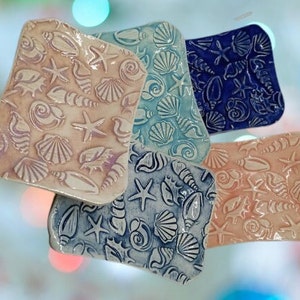 Puede incluir: Una colección de jaboneras de cerámica en varios colores, incluyendo rosa claro, azul y azul marino. Cada plato es rectangular con bordes redondeados y presenta un diseño en relieve de conchas marinas y estrellas de mar. Los platos están dispuestos de forma ligeramente superpuesta.