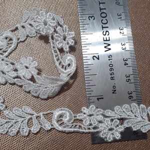 May include: White floral lace trim with a ruler for measurement. The lace features a delicate design of flowers and leaves. The ruler shows measurements in inches, with the lace extending to approximately 3 inches.