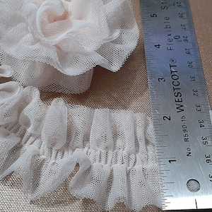 May include: A close-up of a white tulle ruffle trim with a ruler for scale. The trim is approximately 3.5 inches wide and has a delicate, ruffled texture.