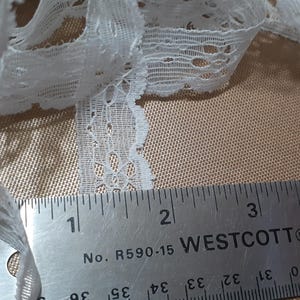 May include: White lace trim with a scalloped edge, measured against a ruler marked in inches. The trim is approximately 1 inch wide.