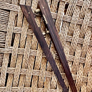 以下が含まれることがあります： 真鍮のアクセントが付いた、2 つの濃い茶色の木製トング。 トングは、ギザギザのエッジを備えたユニークなデザインです。 素朴な美しさを生み出す織りの背景に配置されています。 トングはおそらく食べ物を提供するものです。