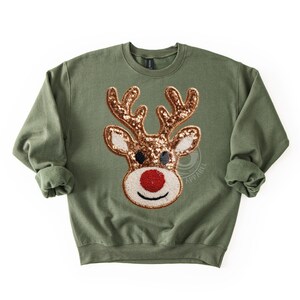 May include: An olive green sweatshirt featuring a sequined reindeer design. The reindeer has brown antlers, black eyes, and a red nose. The sweatshirt has a crew neck and long sleeves, perfect for the holiday season.