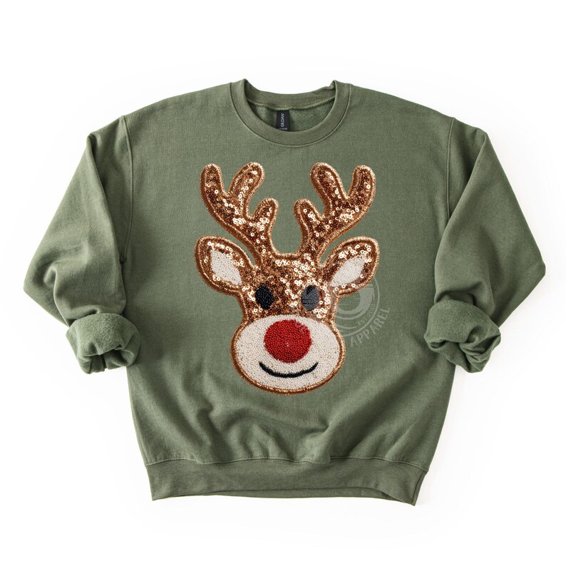 May include: An olive green sweatshirt featuring a sequined reindeer design. The reindeer has brown antlers, black eyes, and a red nose. The sweatshirt has a crew neck and long sleeves, perfect for the holiday season.